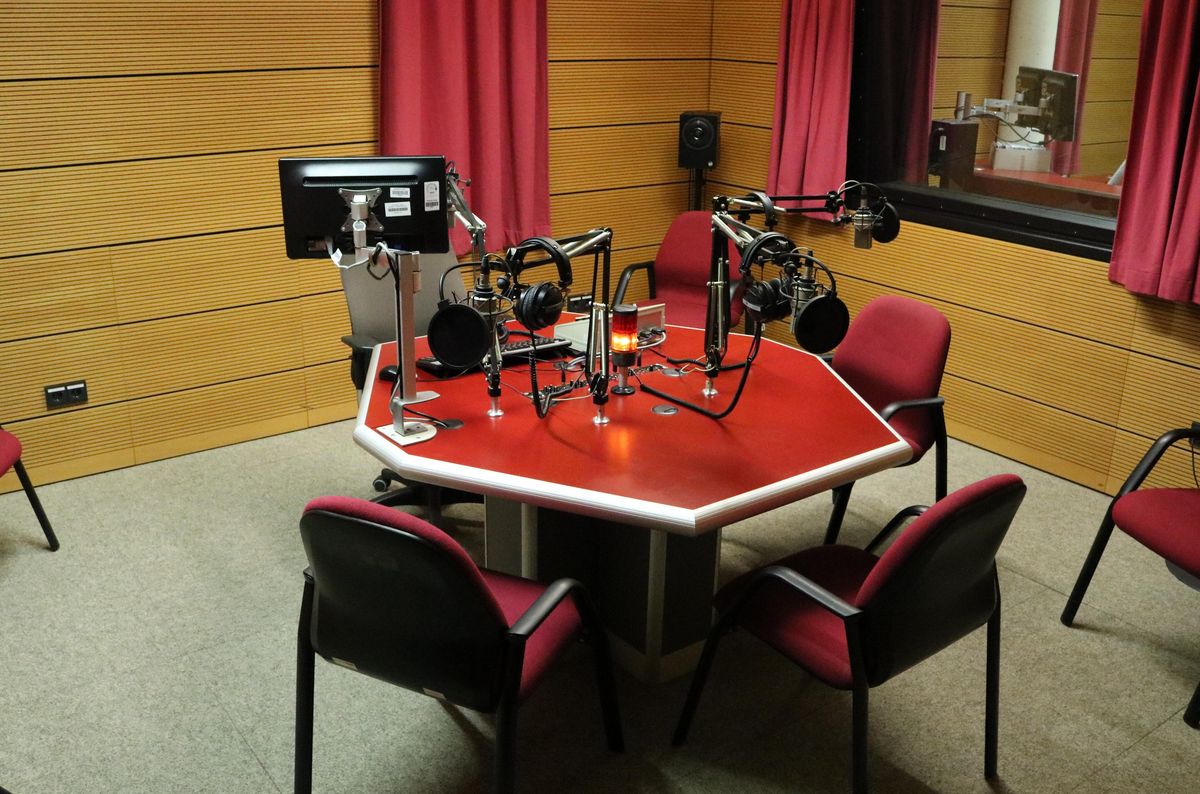 zur Vergrößerungsansicht des Bildes: Studio-Aufnahme: Ein eckiger Tisch in der Mitte mit vier Mikrofonen und Stühlen ringsherum.